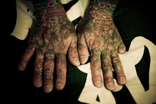 Tätowierte Hände mit fehlender Ziffer. Ein traditionelles japanisches Tattoo, wie es oft von den Yakuza verwendet wird, ist ein sehr altes und zeitaufwendiges Verfahren, bei dem manuell ein Stäbchen mit mehreren scharfen, eingefärbten Nadeln in die Haut gestochen wird. Dies muss in einem genauen Winkel (je nach Hautdicke) und mit einer genauen Geschwindigkeit (120/Minute) geschehen, und diese Fähigkeit besitzen nur traditionelle japanische Tätowierer. Das Ergebnis ist eine Feinheit, eine Farbpalette und ein Muster, das mit der modernen Art des Tätowierens mit einer Maschine nicht möglich ist. Meistertätowierer Hori Sensei lädt Sie ein, er nimmt keine Stammkunden an. Mit ihm dauert die Fertigstellung eines traditionellen japanischen Tattoos etwa 100 Stunden, kann bis zu 10.000 US-Dollar kosten und es muss ein Zeitplan für tägliche oder wöchentliche Besuche erstellt werden. Als Kunde haben Sie nur wenig Einfluss auf die Gestaltung des Tattoos. Hori Sensei bestimmt, was für Sie am besten ist, nachdem er sich die Zeit genommen hat, mit Ihnen zu sprechen und Sie kennenzulernen. Heute leben nur noch wenige traditionelle japanische Tattoo-Experten in Japan. - 2009